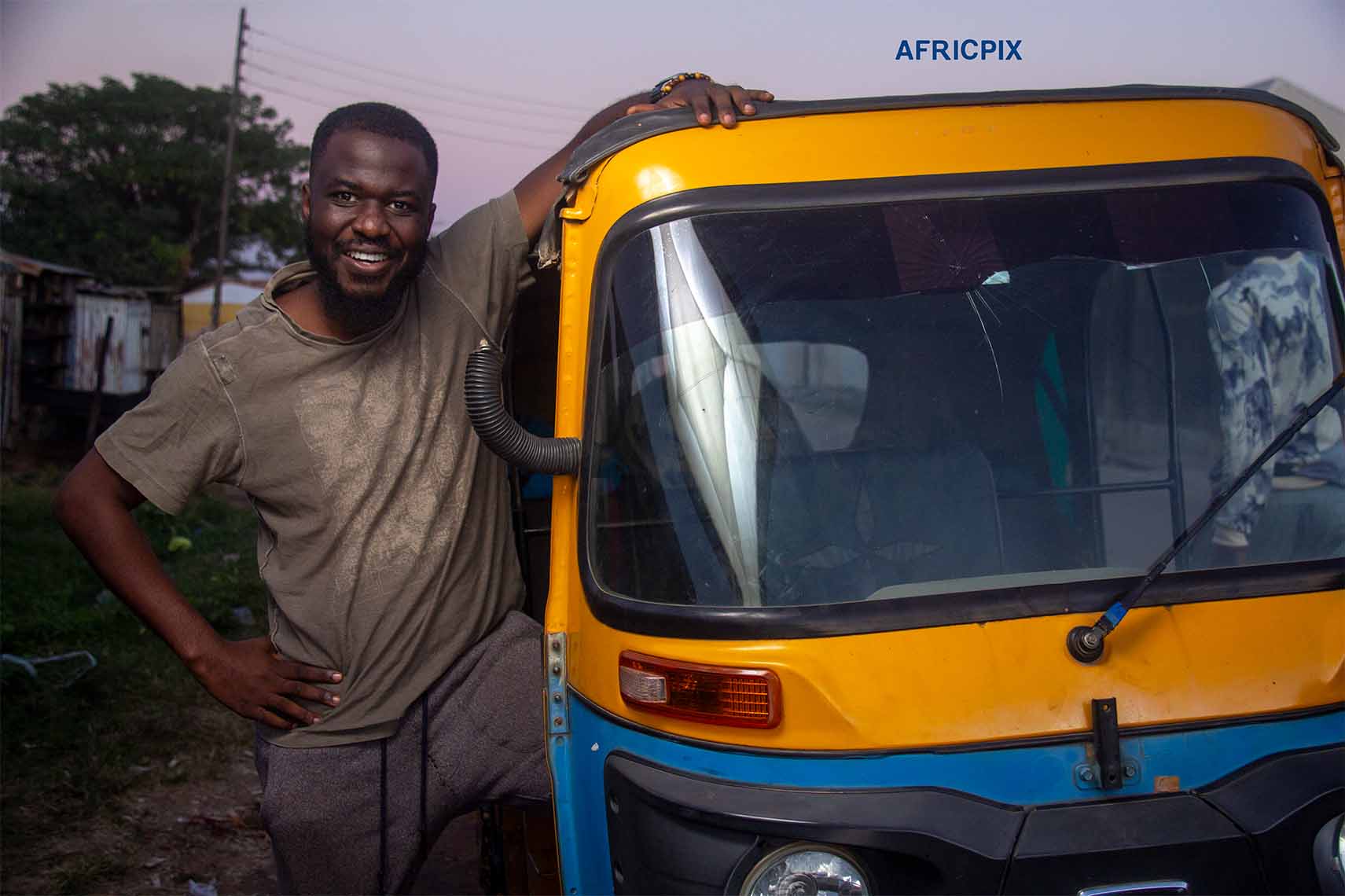 An African man sitting beside a tricycle, keke, maruwa smiling happily